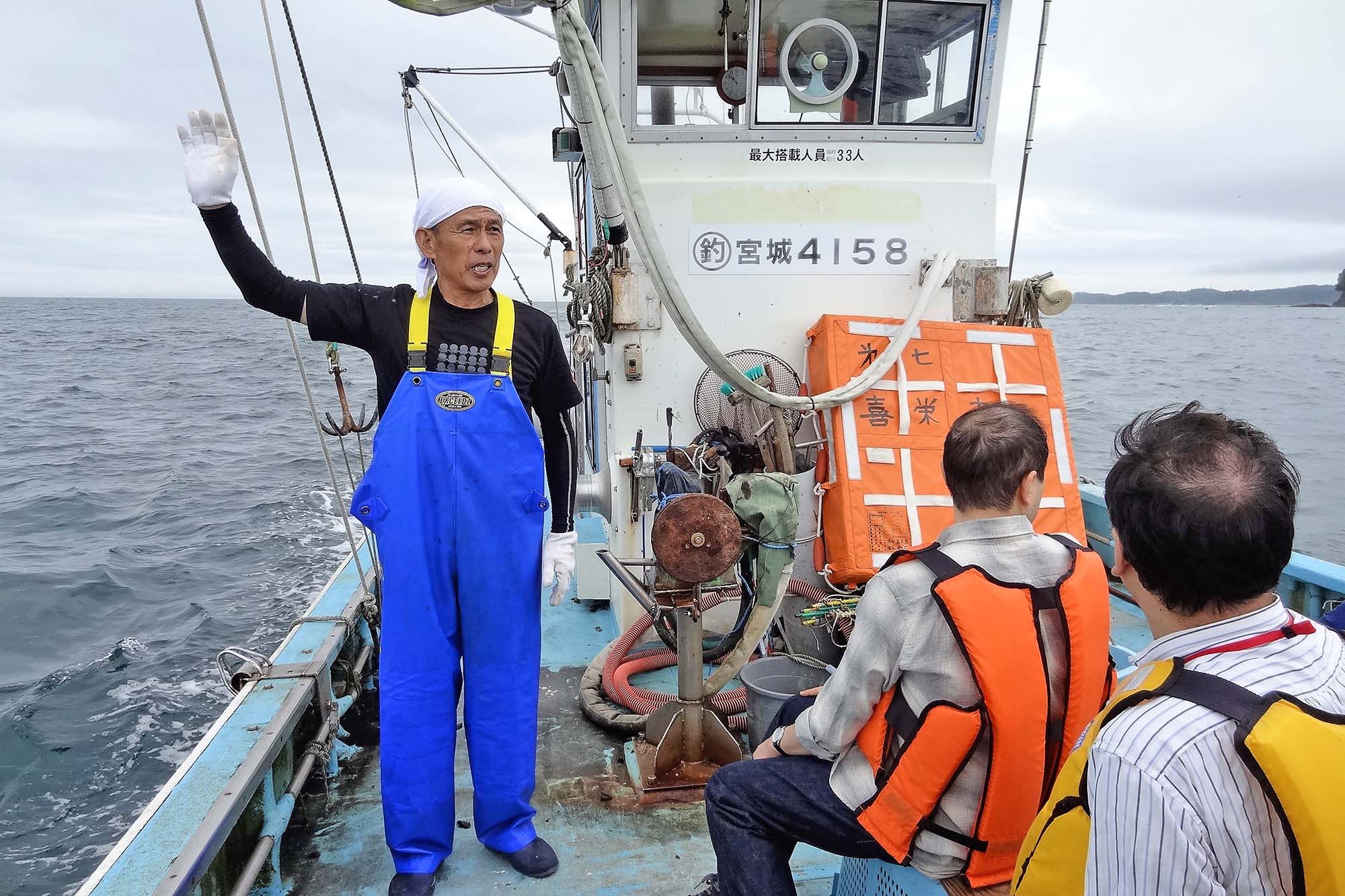 漁船の上で漁師さんが手を挙げて説明し、参加者が救命胴衣を着て話を聞いている写真