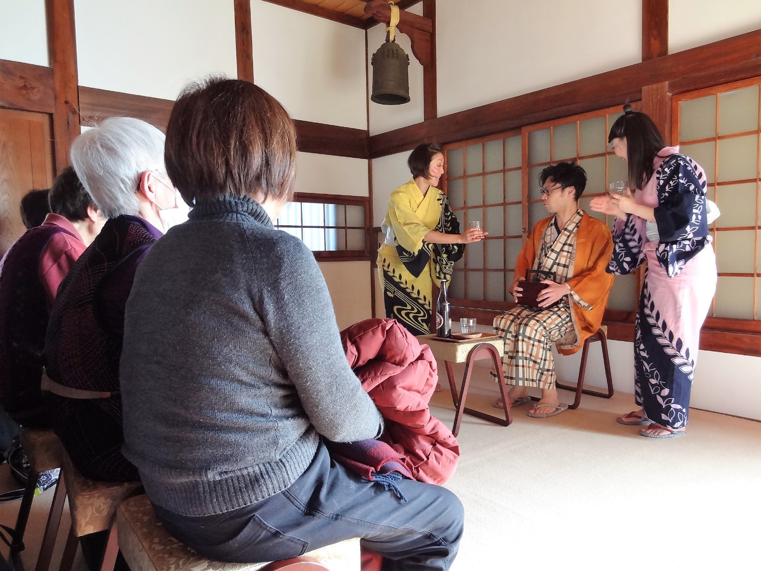 劇を披露している色とりどりの着物を着た3名の方と、椅子に座り観劇している観客達を写した写真