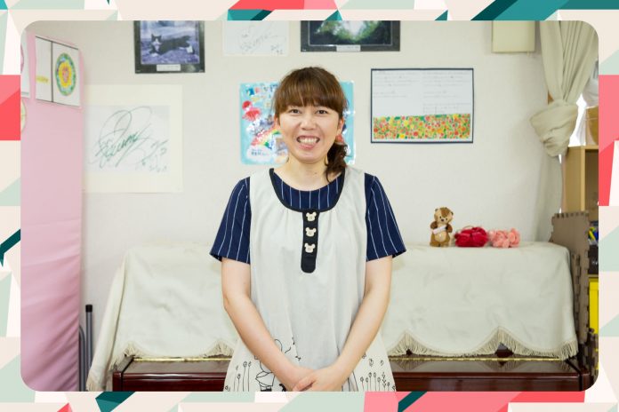 絵や写真などが飾られた壁際に置かれたピアノの前でエプロンを着た高橋しのさんが笑顔でカメラに向かって立っている写真