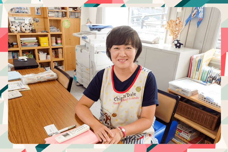 背景には書棚、複合機、本が整理された棚が見える室内で、木製のテーブルの前に座っているチップとデールのキャラクター入りのエプロンを着用した笑顔の女性の先生の写真