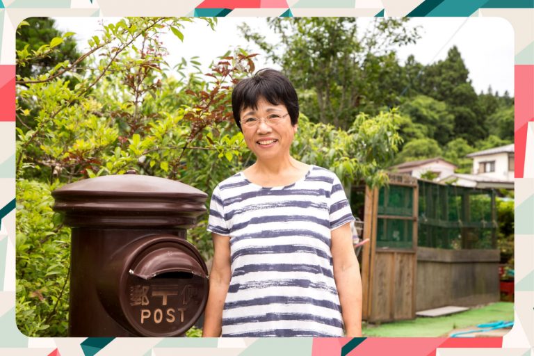 縞模様のシャツを着た女性が、緑豊かな庭と昔ながらの茶色い郵便ポストのそばで微笑んでいる様子をアップで撮影した写真