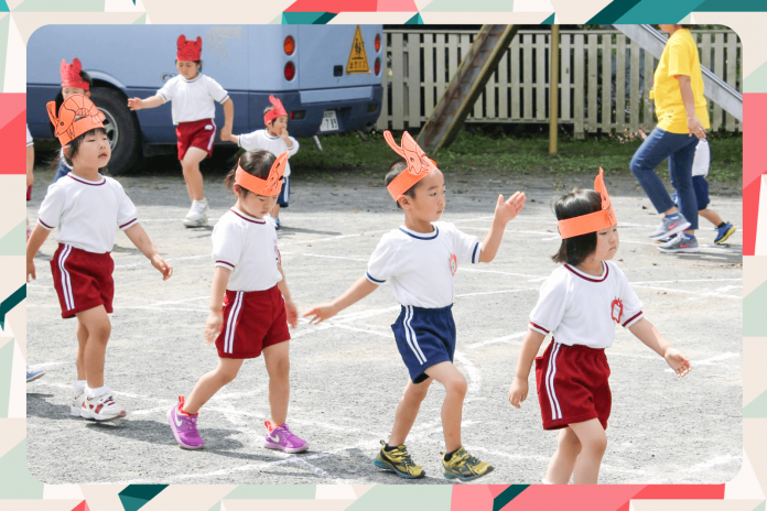 園庭で、数人の園児たちが赤や青の体操服と動物の絵が描かれたオレンジ色の紙の帽子をかぶり、隊列を組んで歩いている様子の写真