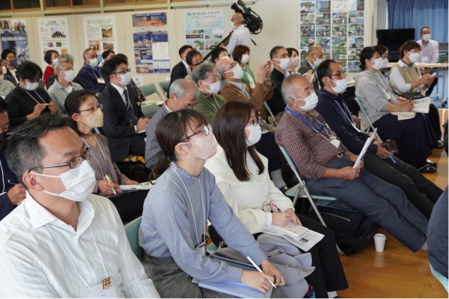 室内の会場で、マスクを着用した多数の参加者たちが椅子に座り、手元の資料を見ながら講演を真剣な表情で聞き入っている様子の写真
