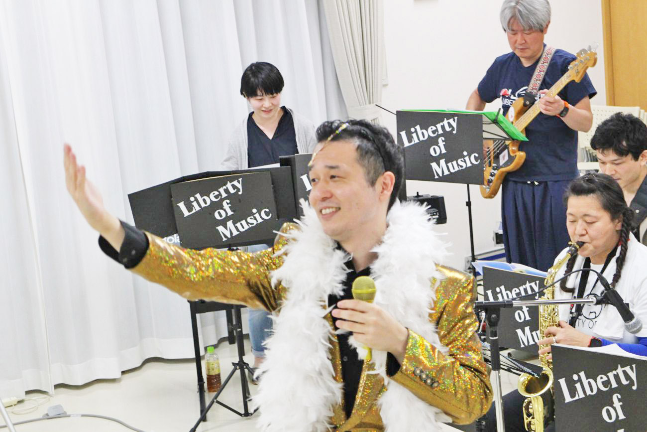 金色のキラキラしたジャケットと白い羽根の飾りを身に着けた男性がマイクを持ち片手を挙げて歌いながら、後ろで数人のバンドメンバーが「Liberty of Music」の譜面台を前に演奏しているライブの写真