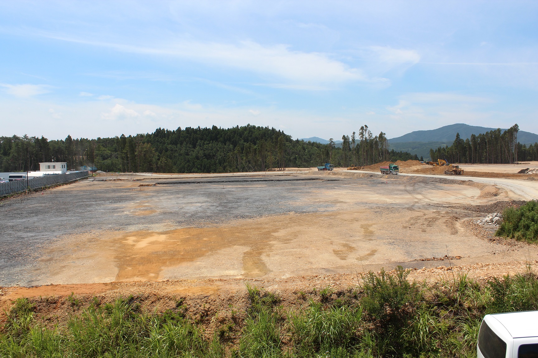 広大な造成地でダンプカーや重機が作業を進めており、背景には緑豊かな森林と山並みが広がっている写真