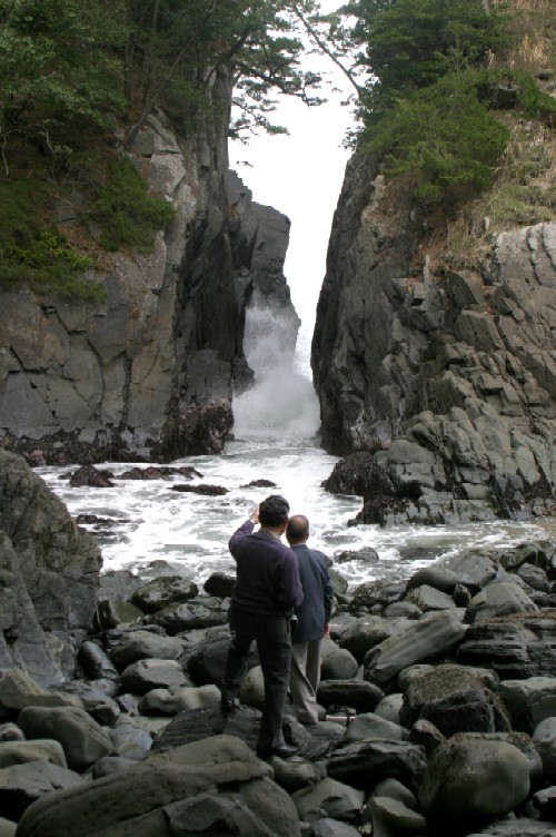 暗い色の岩がごろごろと転がる海岸で、2人の男性が波間に向かって立つ大きな岩と岩の裂け目から激しく波が打ちつける場所を眺めている、自然の力の強さを感じる風景写真