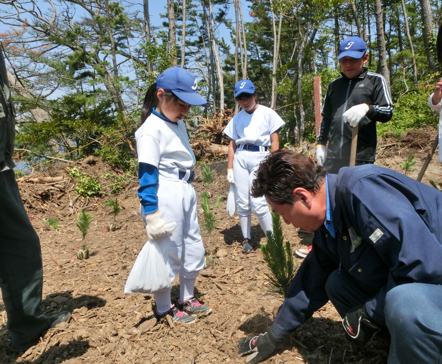 松の苗木が植えられている場所で、植樹の手順を説明している男性の周りで野球のユニフォームを着た女の子や男性が話を聞いている様子の写真