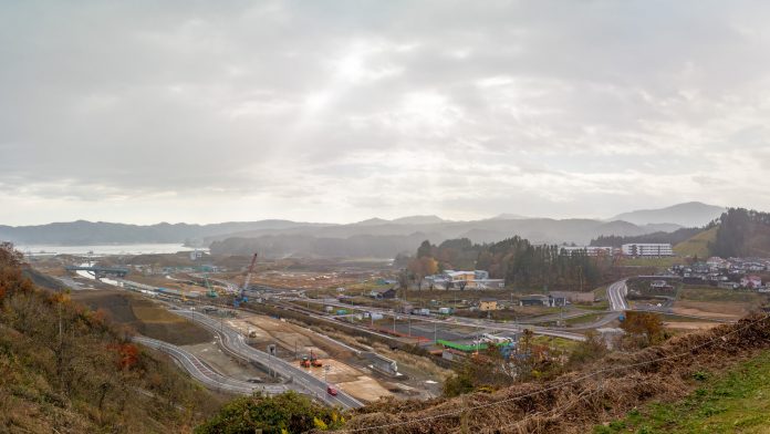 複数の道路があり、一部は工事中で、建設車両や機材が点在し、周囲には丘や木々、点在する建物が見られ、背景には山々が広がっており空は雲が広がって曇っている進行中の建設作業現場を写した写真