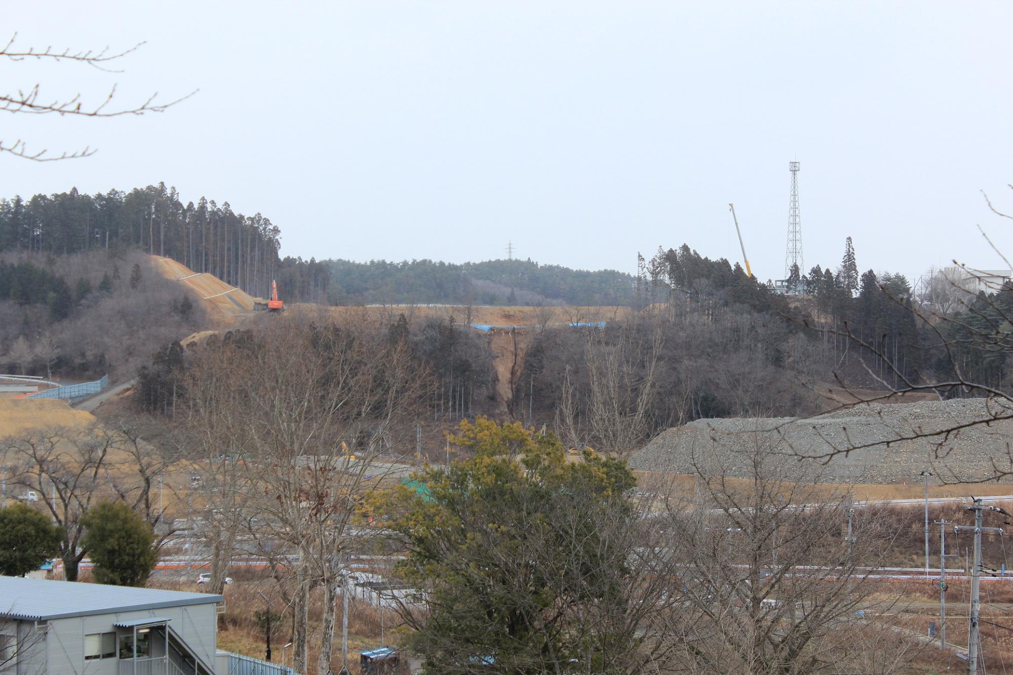 周りの木々の葉がすっかり落ち、奥の様子がよく見える造成地に1台のオレンジ色のショベルカーやクレーンのブーム部分が伸びている様子が伺える写真