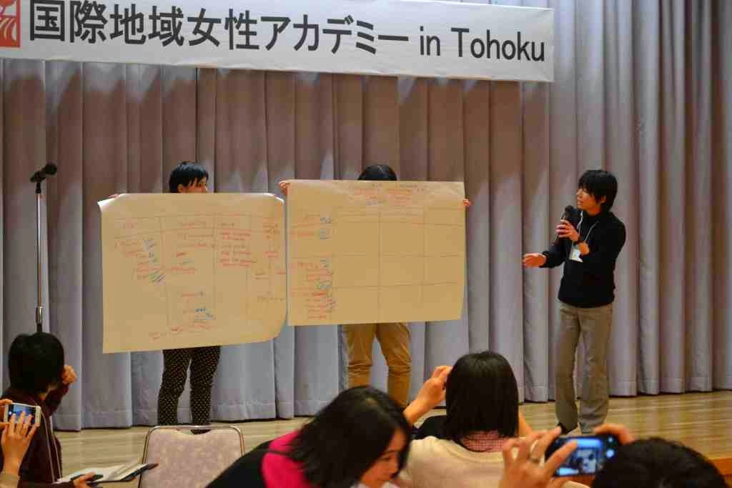 2枚の資料を持っている人と、舞台の上で説明をしている女性の写真