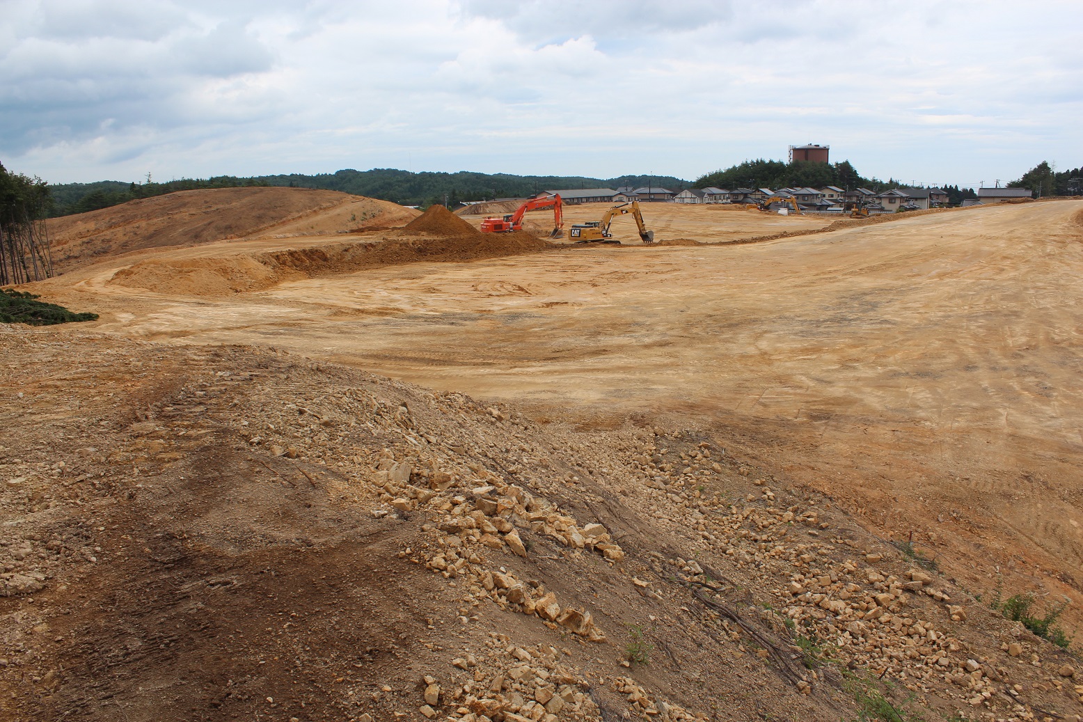 高台に造成された広い土地に重機が複数稼働しており、整地や基礎工事が進められる中、遠景には山々が連なり、復興の一環としての住宅建設準備の様子がうかがえる写真