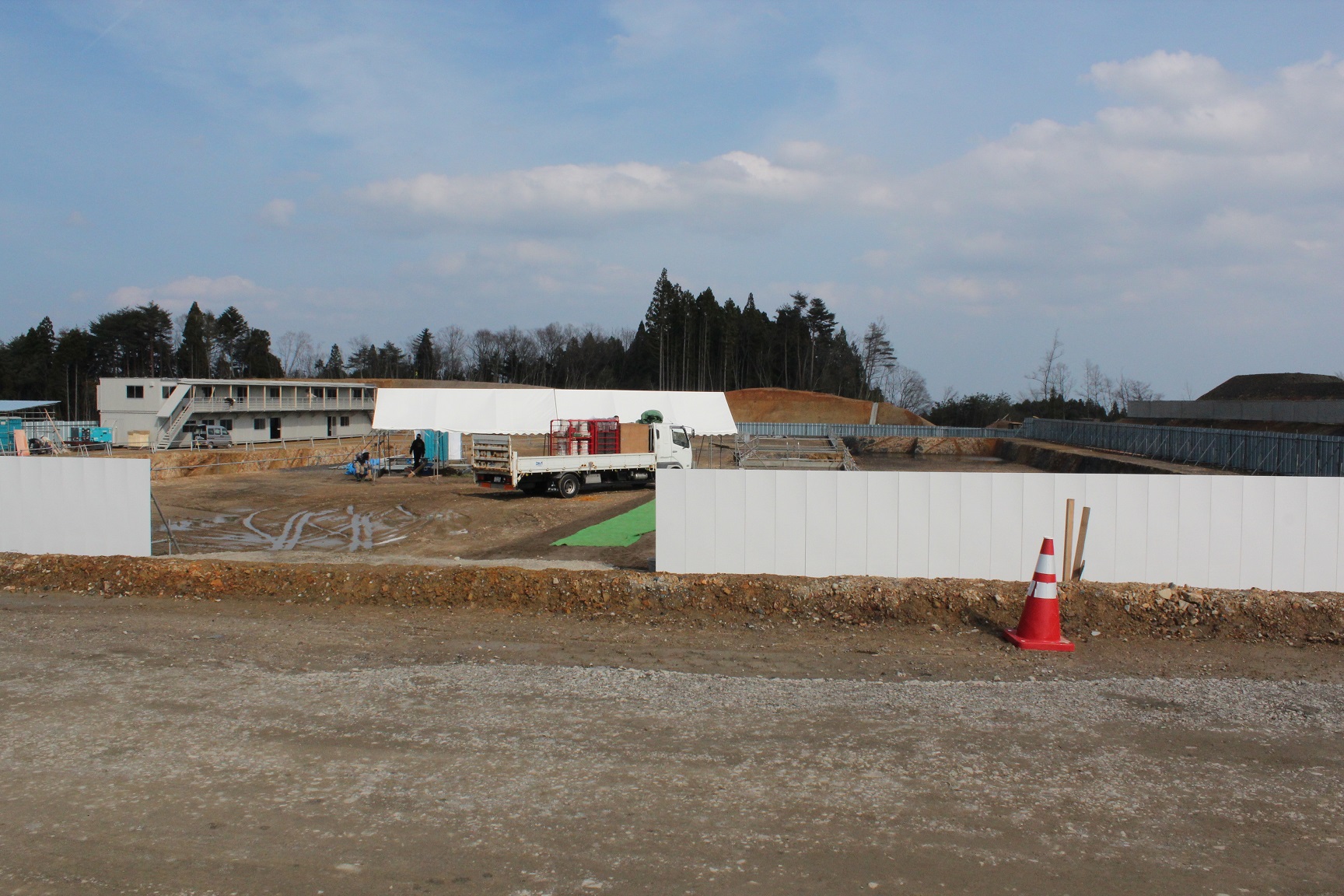 白いフェンスのようなもので囲まれた建設現場の様子を示しており、中央にはトラックが停まっていて、その周囲には工事用の資材や機材が散らばっており、背景には木々が立ち並んでいる写真