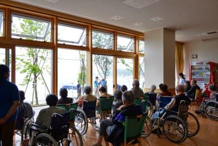 大きな窓から見える外の景色を見ながら、椅子や車椅子に座った多くの入居者が並んで植樹している様子を眺めている写真