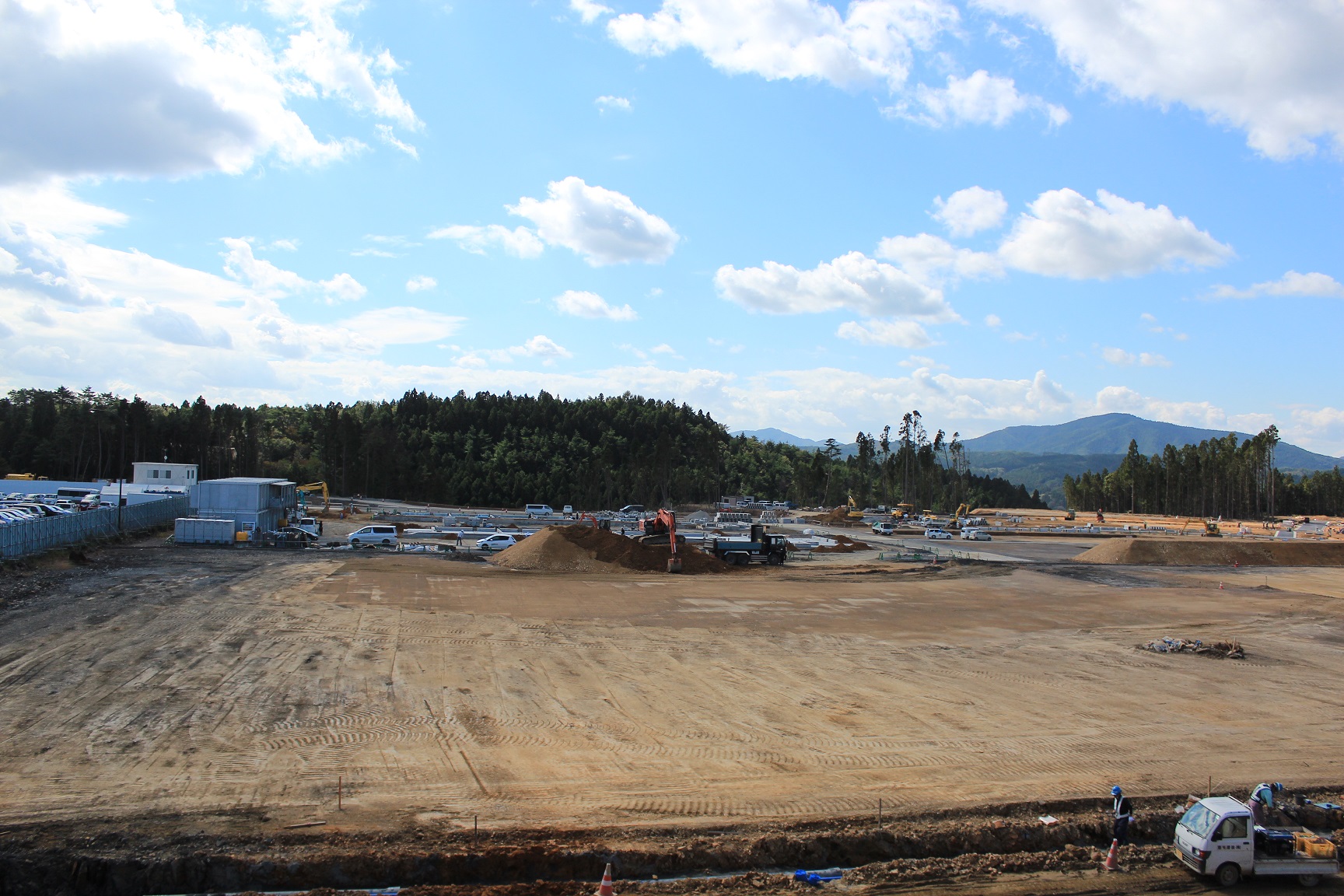 晴天のもと、広大な造成地で複数の重機や作業車が稼働し、中央ではショベルカーが土を積み上げ、手前では作業員が地面を整備している様子の写真