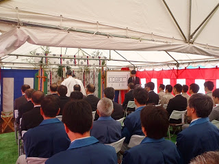 紅白幕が張られたテントの中で、参加者たちが椅子に座り前方の神棚で神職の男性が神事を執り行っている安全祈願祭の様子の写真