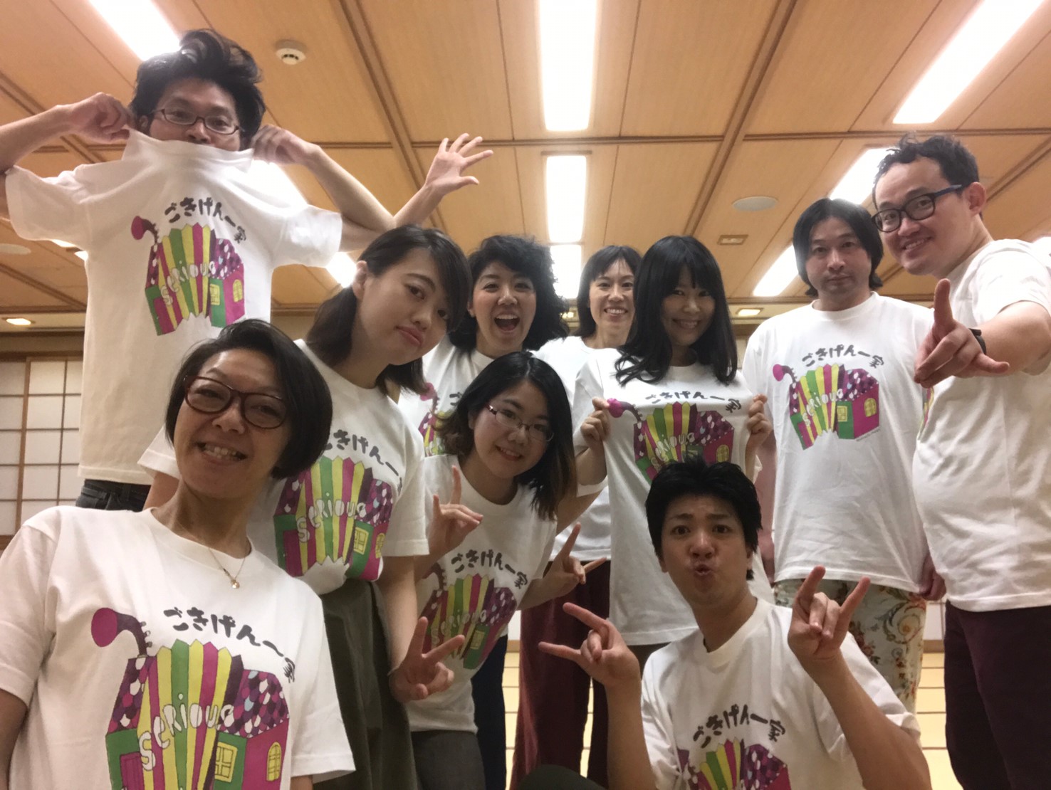「ごきげん一家」と書かれたお揃いの白いTシャツを着た10人の団員が、室内でピースやポーズを決めて並んでいる集合写真