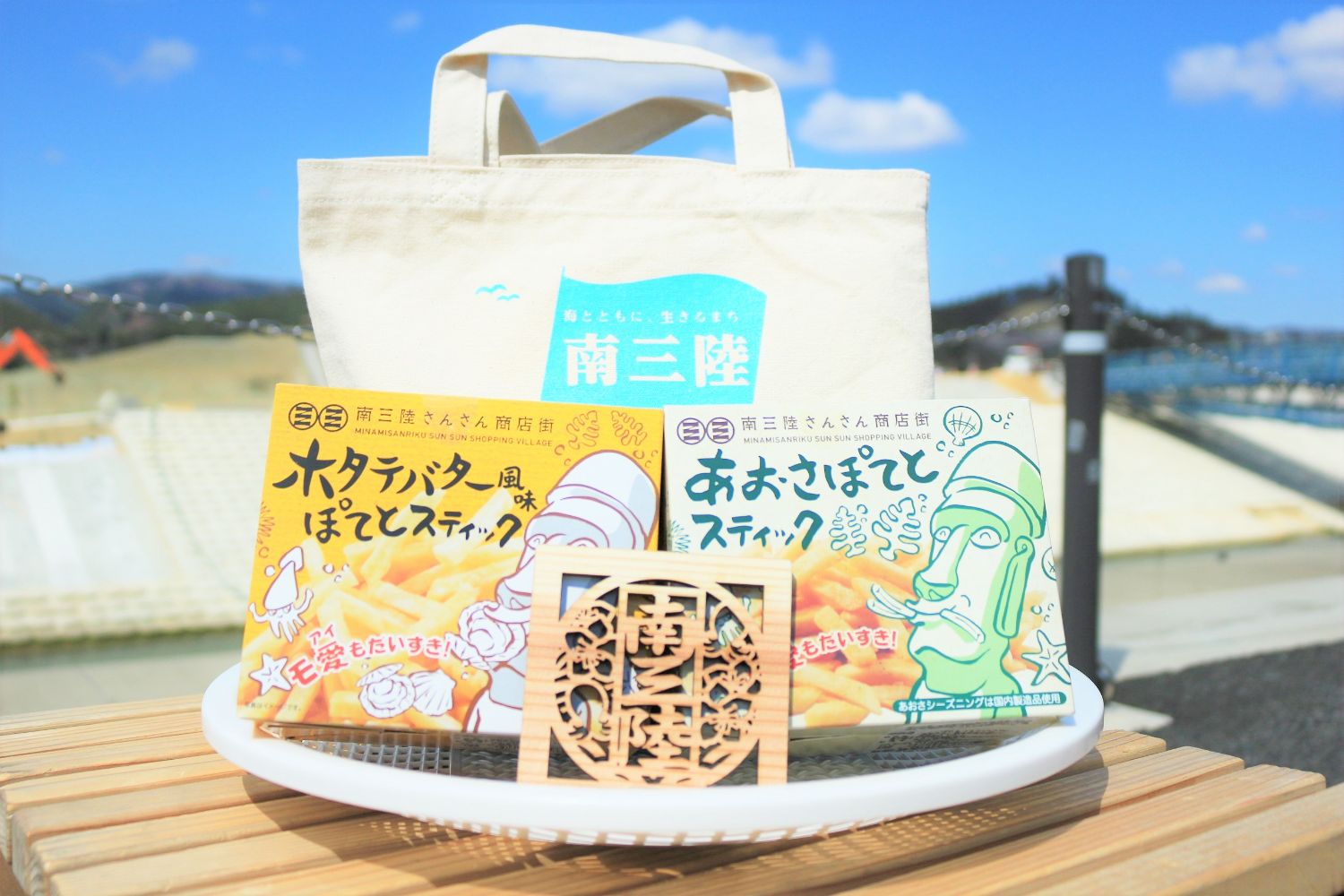 三陸の観光地で販売されているモアイキャラクターが描かれたホタテバター味とアオサ味のスティック菓子がトートバッグと一緒に屋外テーブルに置かれている写真