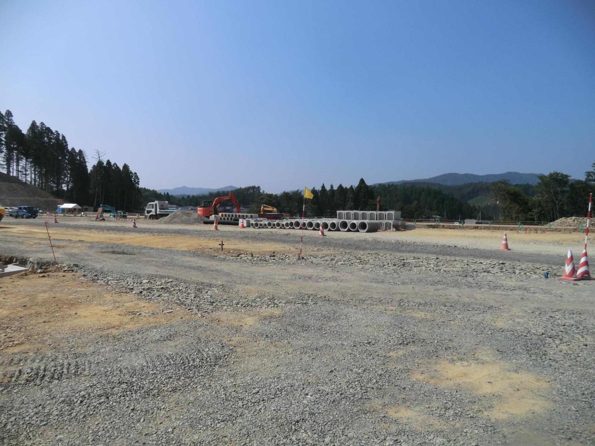 山々に囲まれた広大な造成地で、地面には砕石が敷かれ、中央には整然と並べられた複数の大型コンクリート管と重機、トラックが配置されている様子が写る写真
