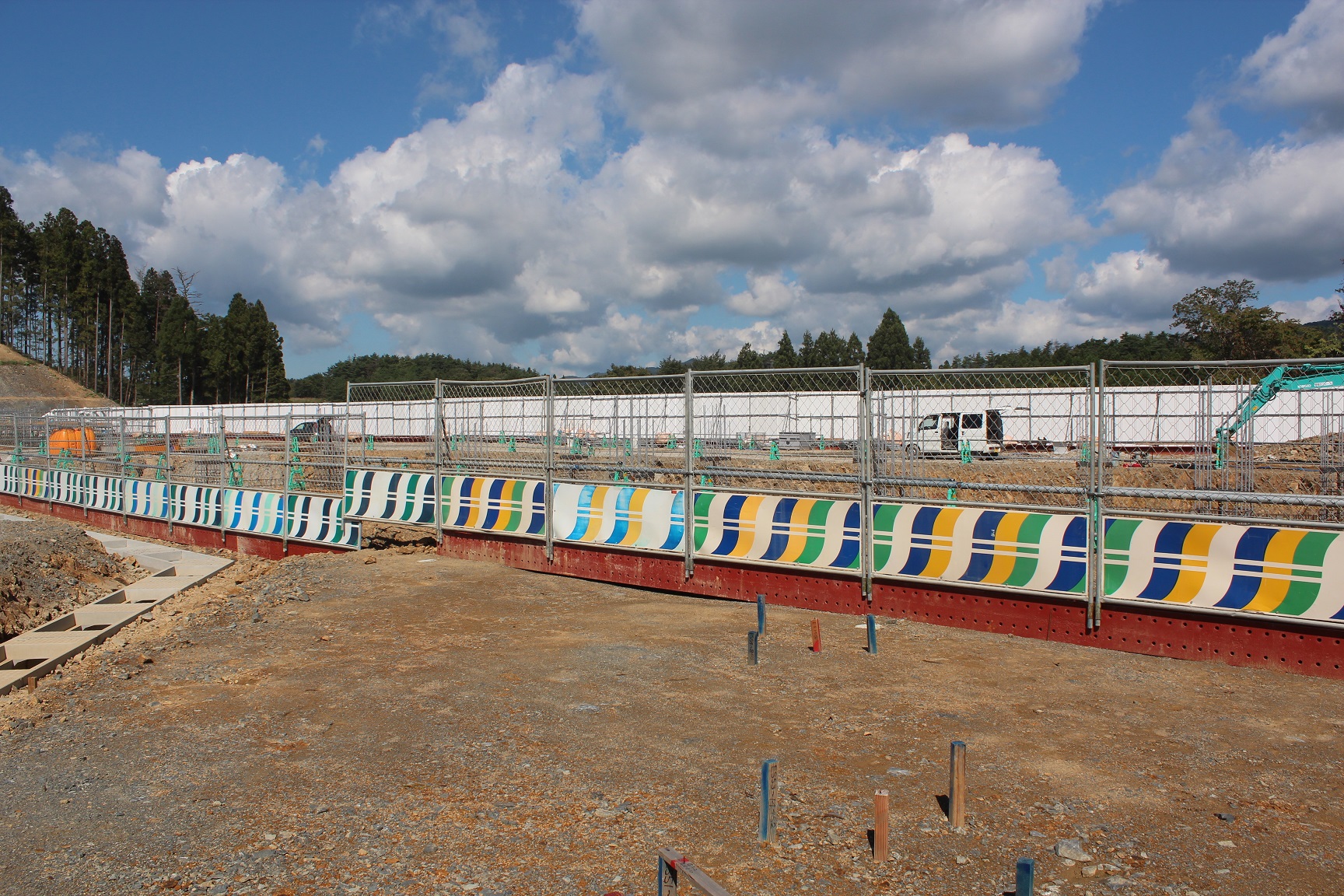 青空と白い雲の下、カラフルな模様のフェンスで囲まれた建設現場で、基礎工事が進められ、奥には重機やトラックが並ぶ様子が見える写真