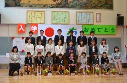 国旗と町旗に「入学おめでとう」と書かれた室内で、8人の新入生と保護者の方々、両脇に2人の女性教師が並んで座っている記念写真