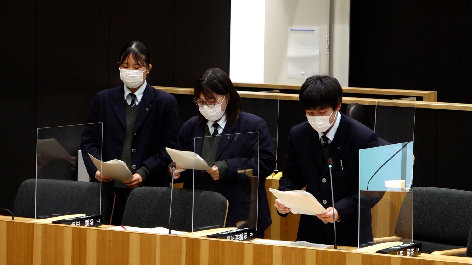 議場で3人の生徒が立ち上がり、、手元の資料を見ながら真剣な表情で発表に臨んでいる写真
