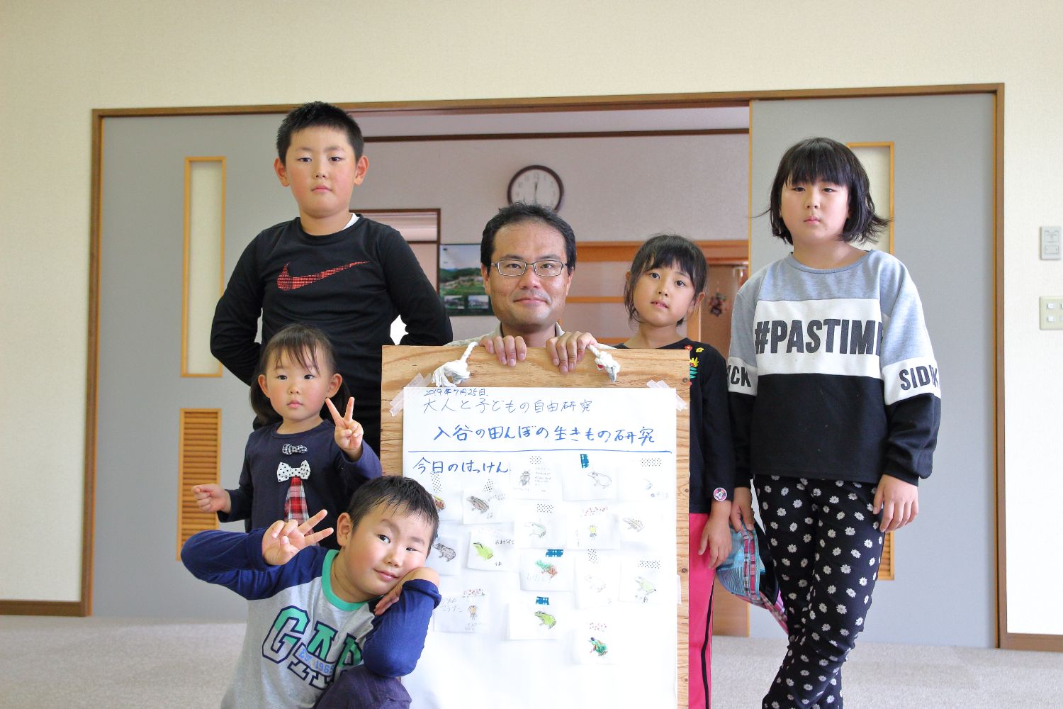 数人の子どもたちと講師の男性が、田んぼで見つけた生きものの絵を貼った大きな発表ボードを囲んで集合写真を撮っている様子の写真