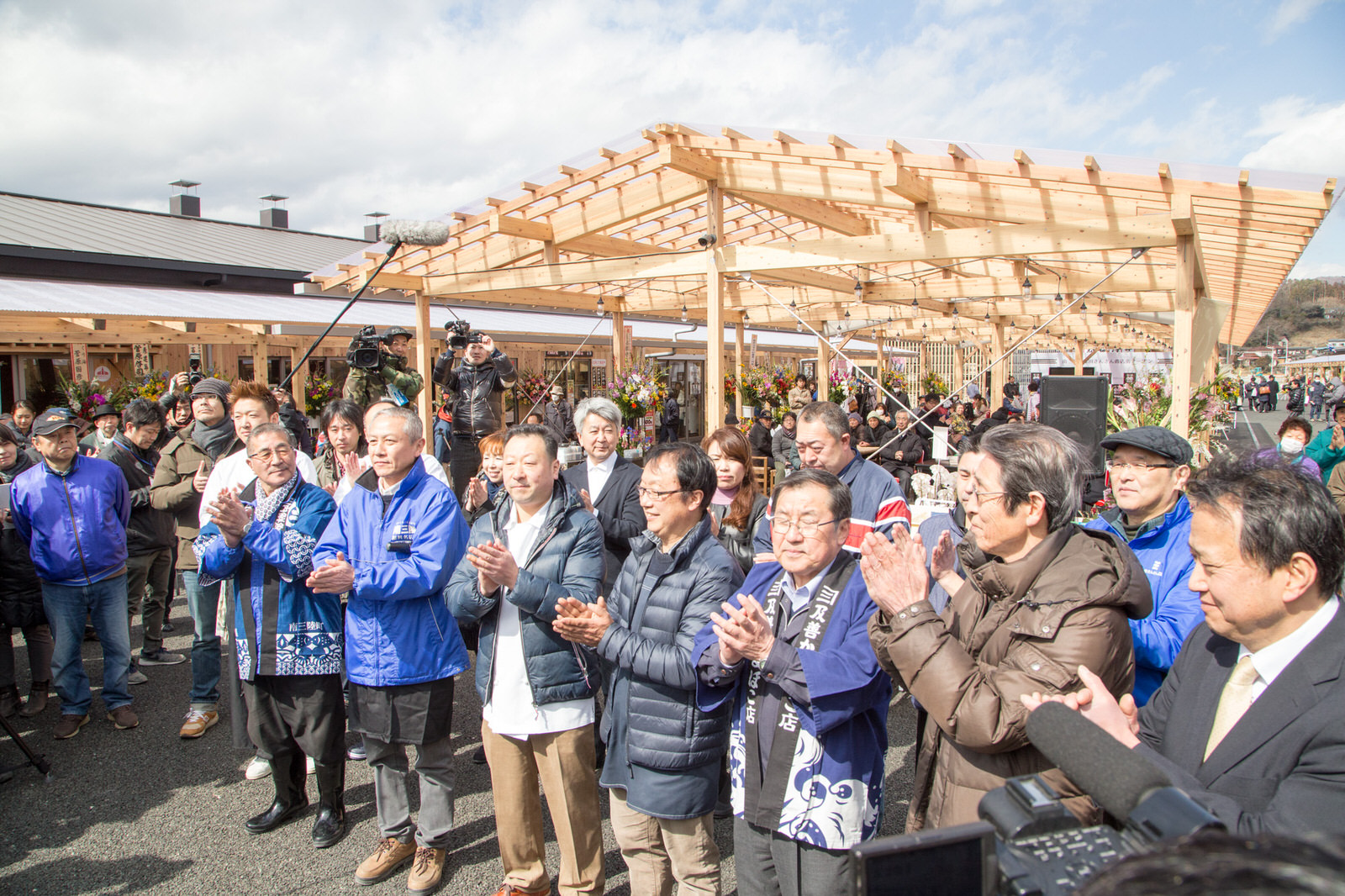 周囲にはカメラマンや多くの人々が屋外の木造建物の前に集まり、中央にはスーツや防寒着を着た関係者が並んで拍手をしているオープンを宣言している様子の写真