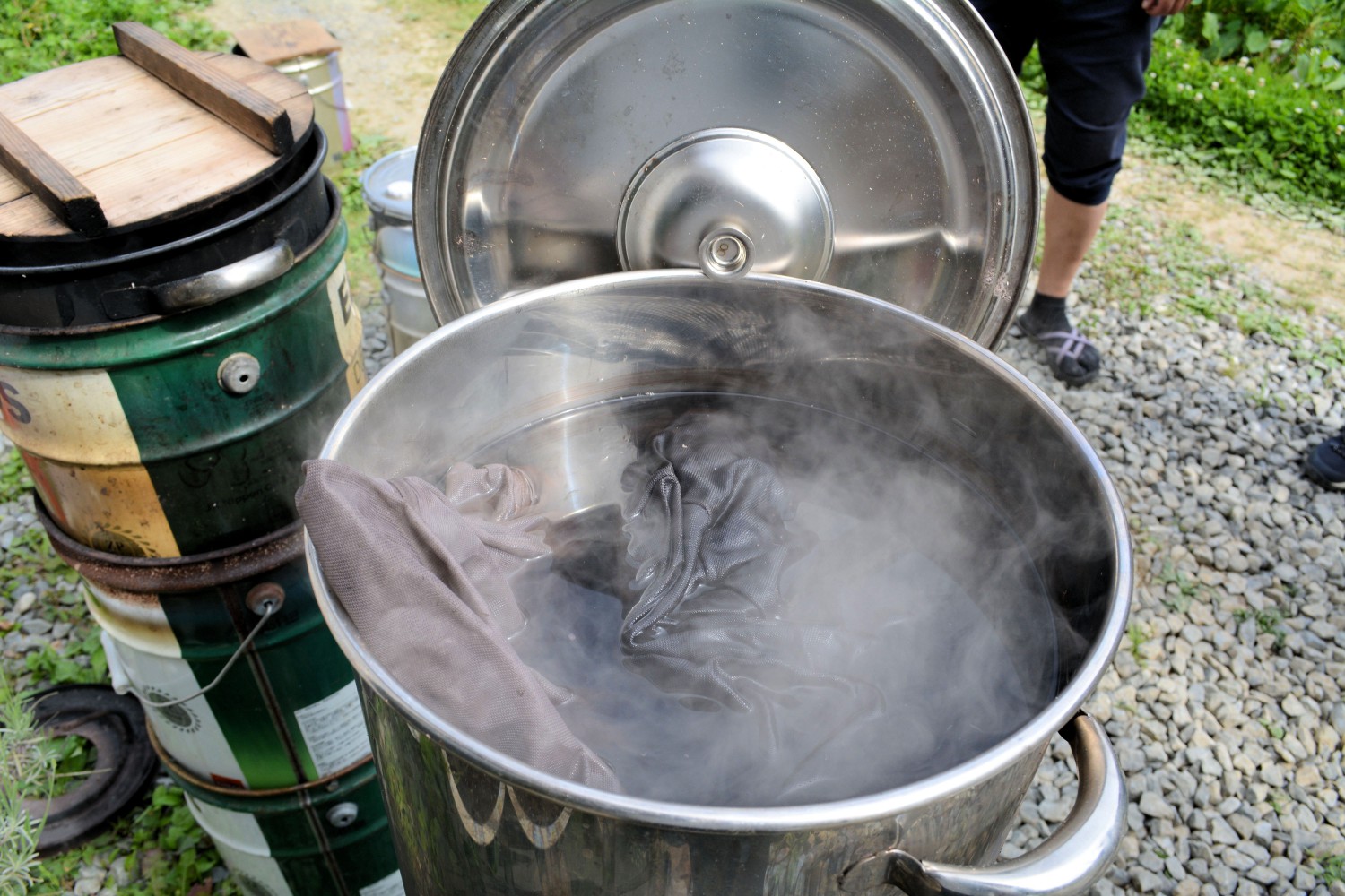 石が敷き詰められた屋外で、蒸気をあげる大きな金属鍋の中に羊毛が入ったネットが入っている写真