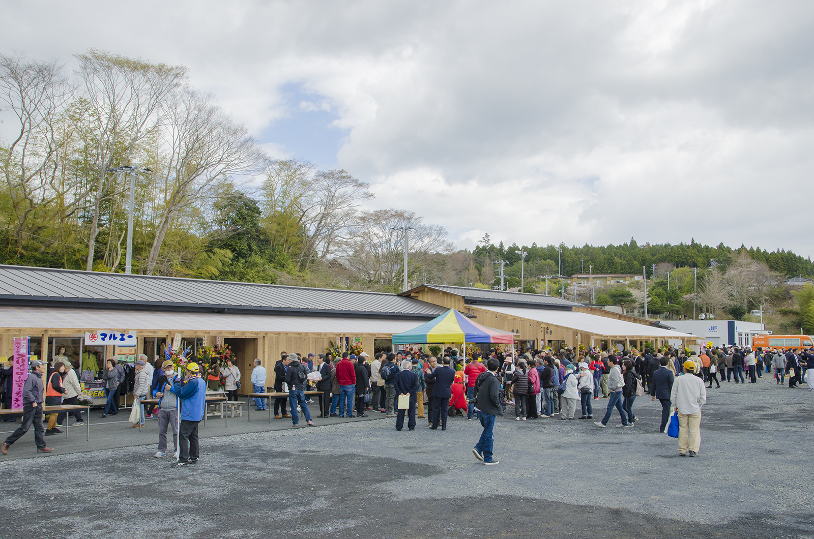 林を背後に建つ新しくできた商店街に多くの人々が訪れ、テントの周辺にたくさんの人が集まっている写真