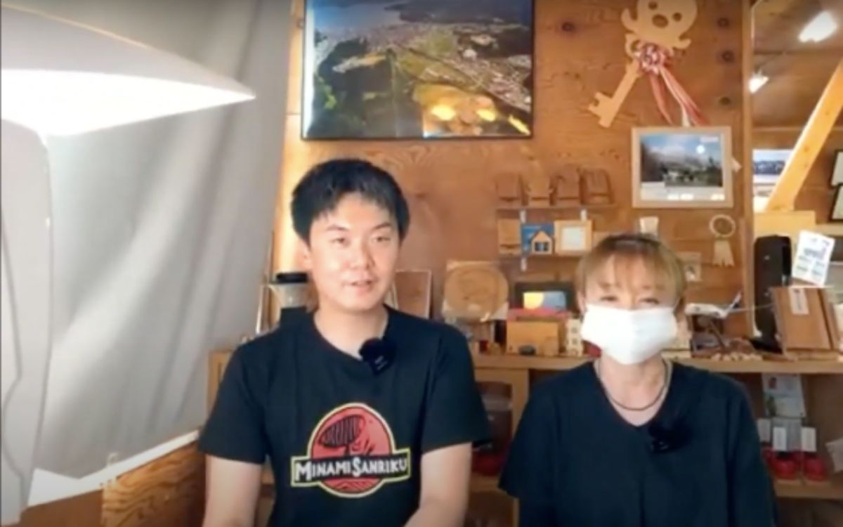 背後の棚には木工品や写真、地図、飾りなどが整然と並んでいる室内で、男性と白いマスクをした女性が並んで座っている写真