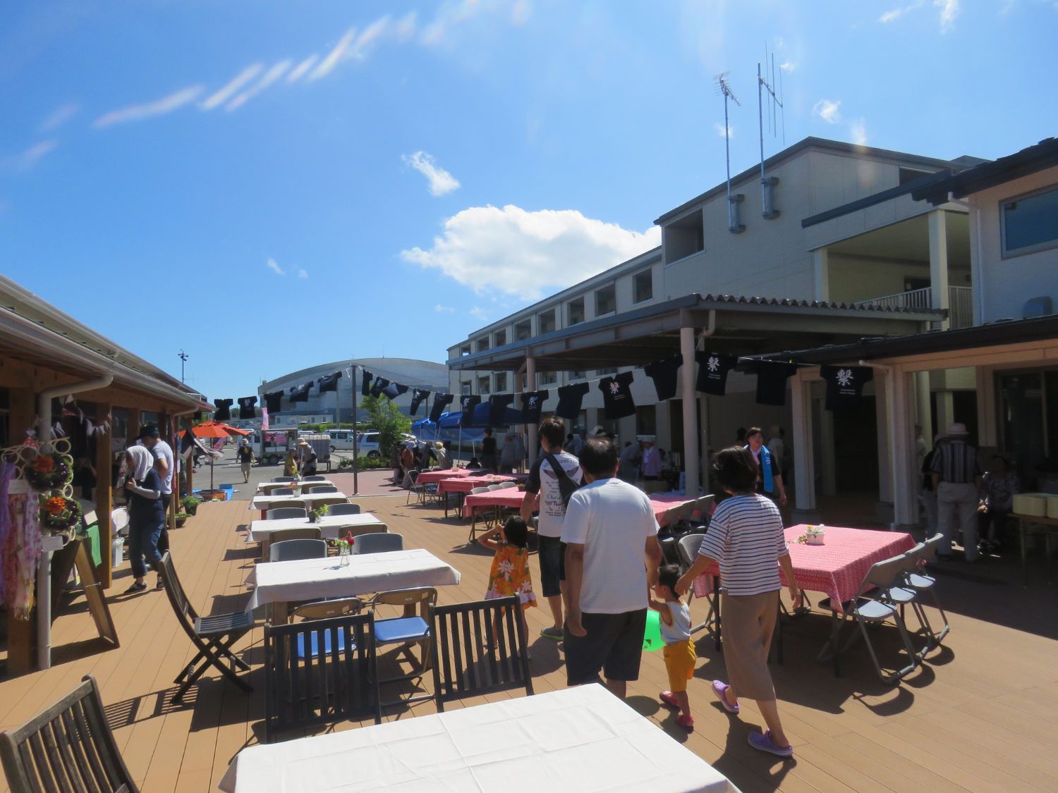 青空の下、ウッドデッキに白や赤のテーブルクロスがかけられたテーブルが並び、多くの人々が散策したり座ったりして賑わいを見せる中、頭上には「繋」などの文字が入った紺色のTシャツがロープに吊るされて風に揺れている写真