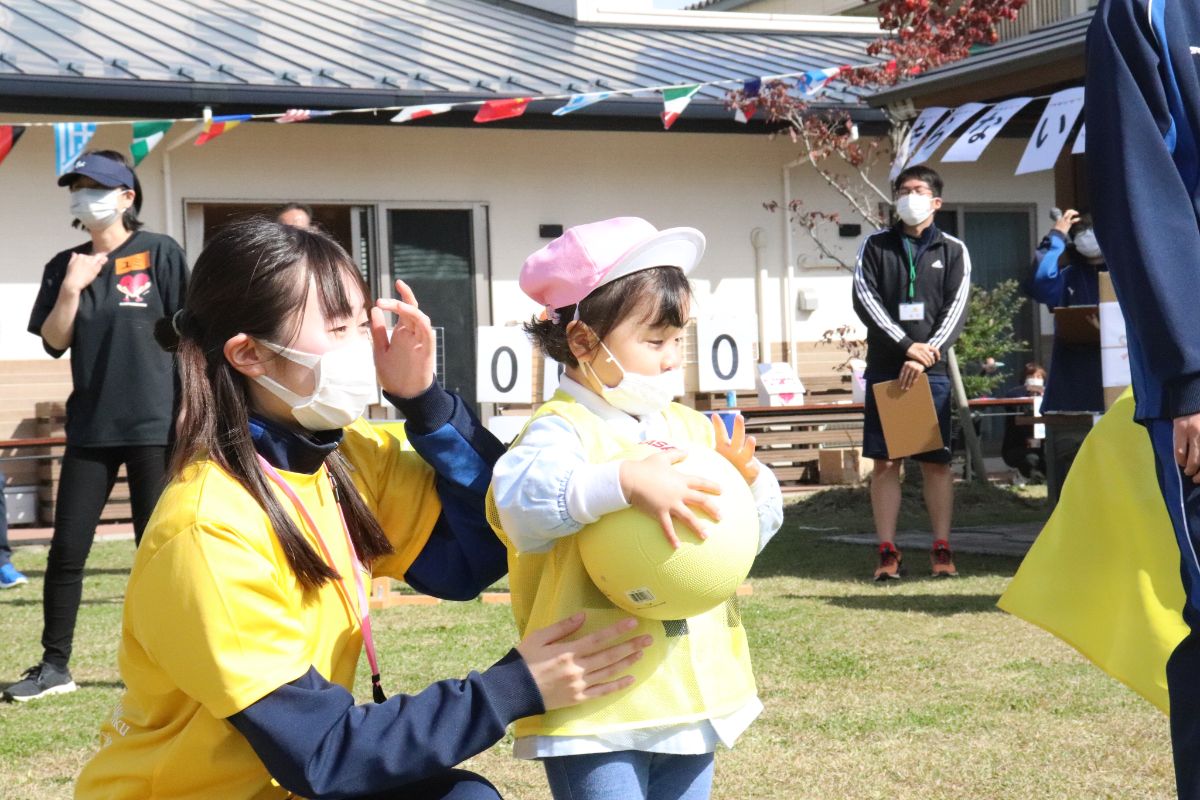 黄色のビブスとピンクの帽子をかぶった幼児がボールを持ち、女子生徒が寄り添って運動会の種目に挑んでいる写真