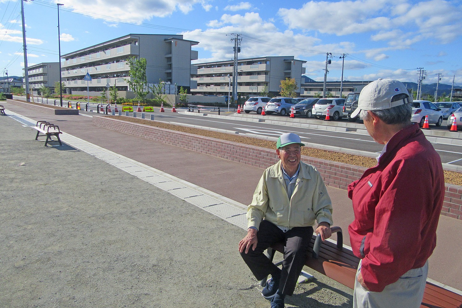 団地や駐車場が見える歩道沿いの屋外のベンチに座る高齢男性と、横に立った男性が笑顔で会話している写真