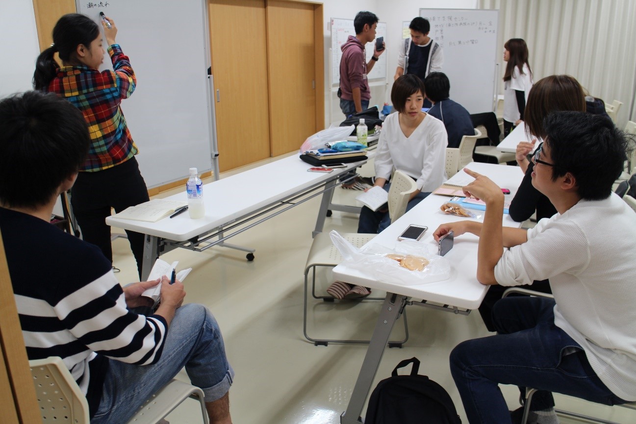 教室の中で、数人の学生が立ち上がってホワイトボードの前に集まり、書き込みをしながら議論や作業を進めている様子が写っている写真