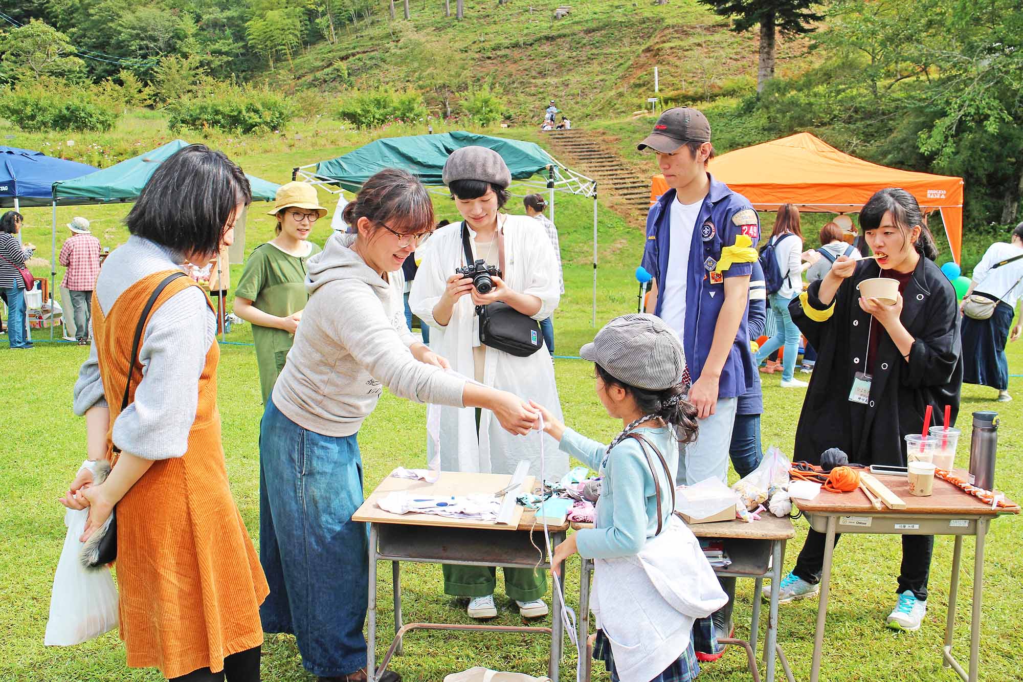 野外の芝生広場に設けられたブースで、スタッフの女性が編み糸を手渡しながら子どもに笑顔で接しており、周囲に参加者の皆さんが数名集まっている様子の写真