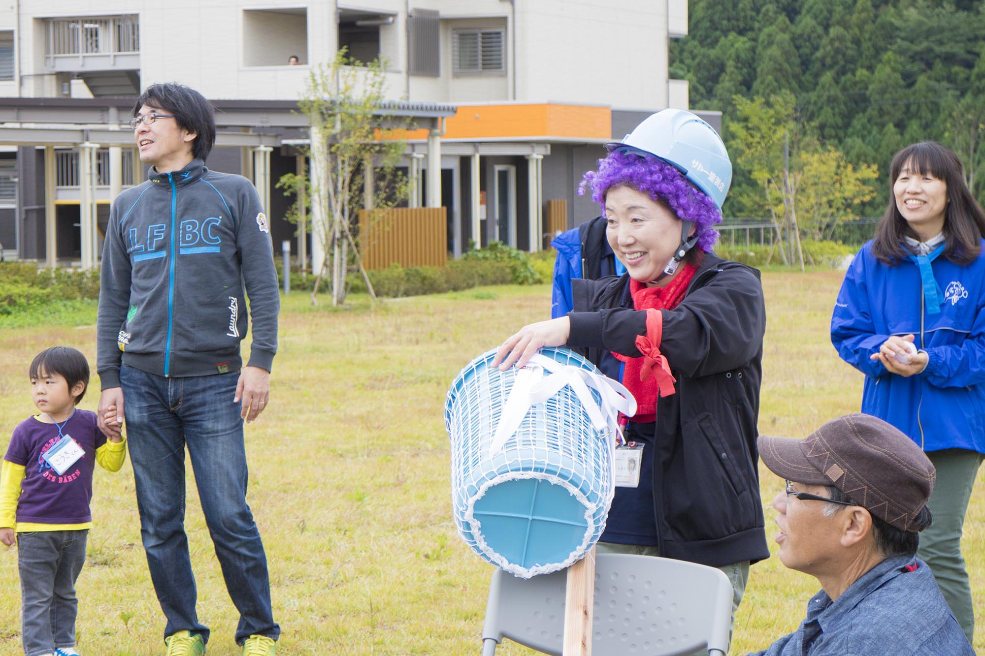 紫のカツラと青いヘルメットをかぶった女性が笑顔でリボンのついた水色のバケツを持っている写真