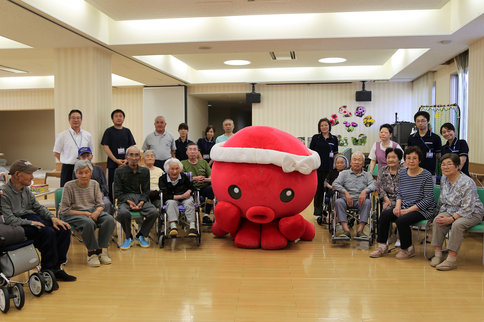 赤いタコのキャラクター「オクトパス君」が、老人保健施設で車椅子に座る高齢者や職員たちに囲まれ、中央に座って記念撮影している写真