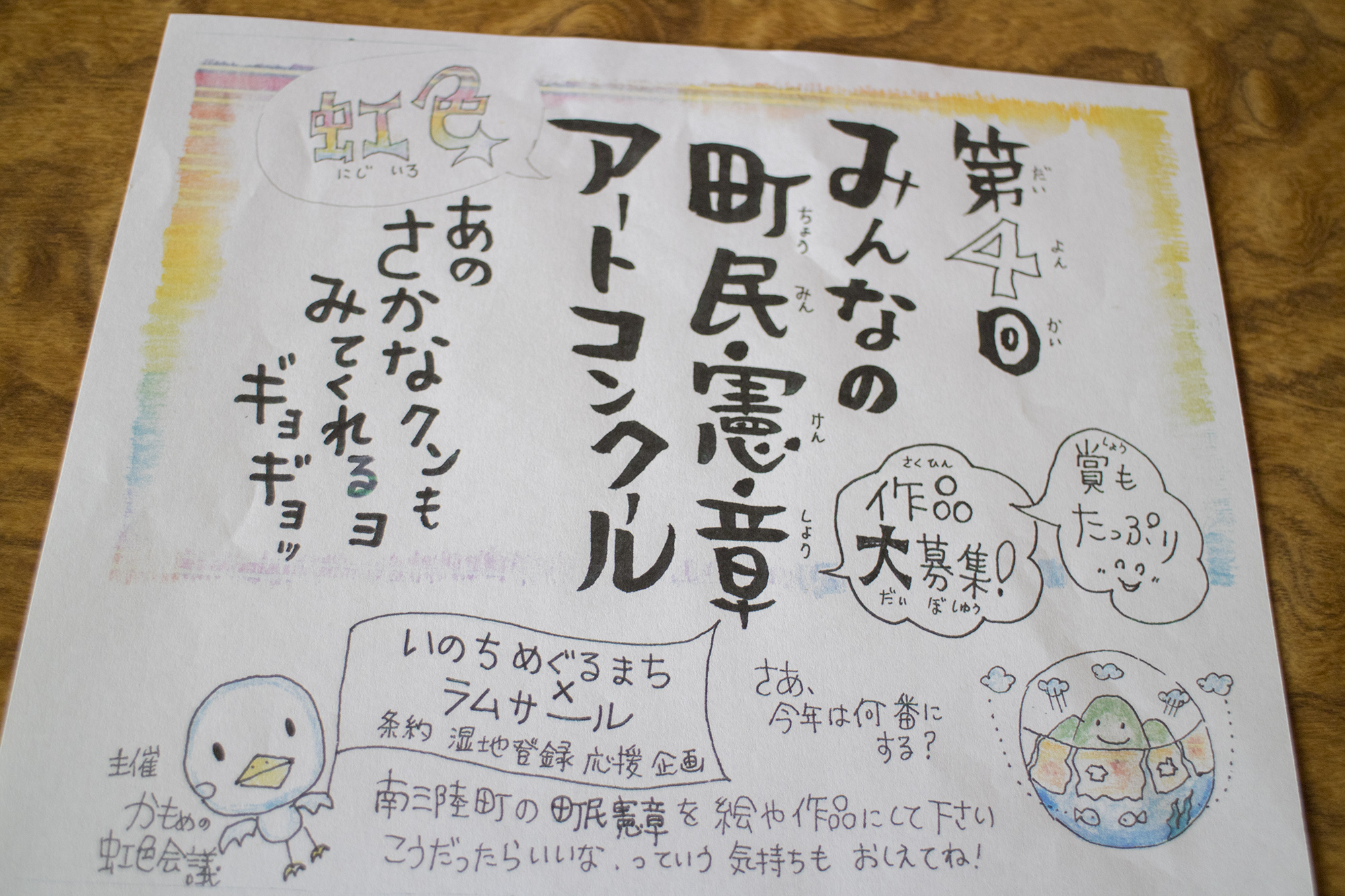 「第4回みんなの町民憲章アートコンクールの作品募集チラシで、手書き風の文字やカラフルな虹の枠、キャラクターのイラストなどが賑やかに描かれている写真