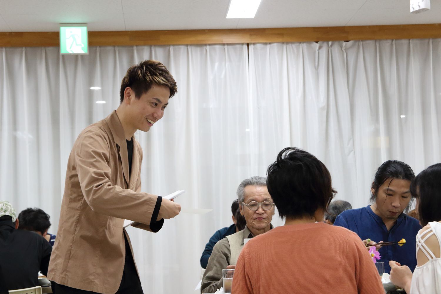 薄いベージュ色のジャケットを着た企画者の岩田さんが、お披露目会で食事をしている参加者の方々に用紙を配布している写真