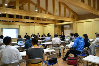 会場の前方に設置されたモニターには映像が映されており、関係者が前方に並んで座る中、多くの参加者が長机に着席して、配布された資料に目を通しながら真剣に話を聞いている様子の写真