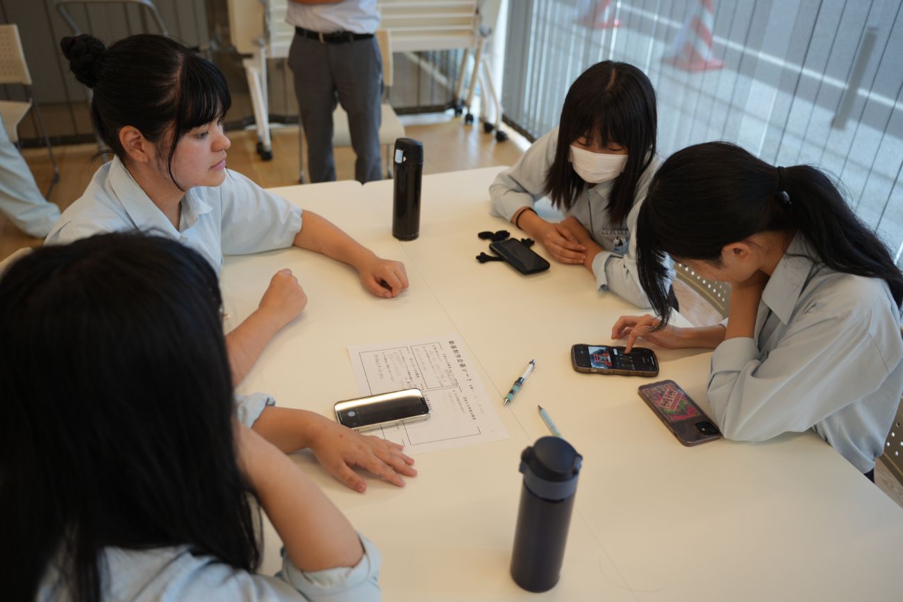 高校生4人がテーブルを囲み、動画をスマートフォンで確認しながら話し合っている様子の写真