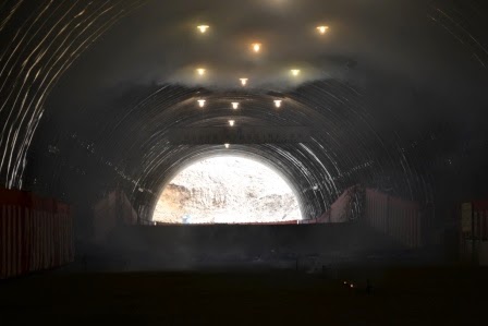 曲線状の天井を持つ大きなトンネルの内部が写されており、天井には複数の照明が設置され、内部には煙が漂っていて、奥には外の自然光が差し込む出口が見えているトンネル内の写真