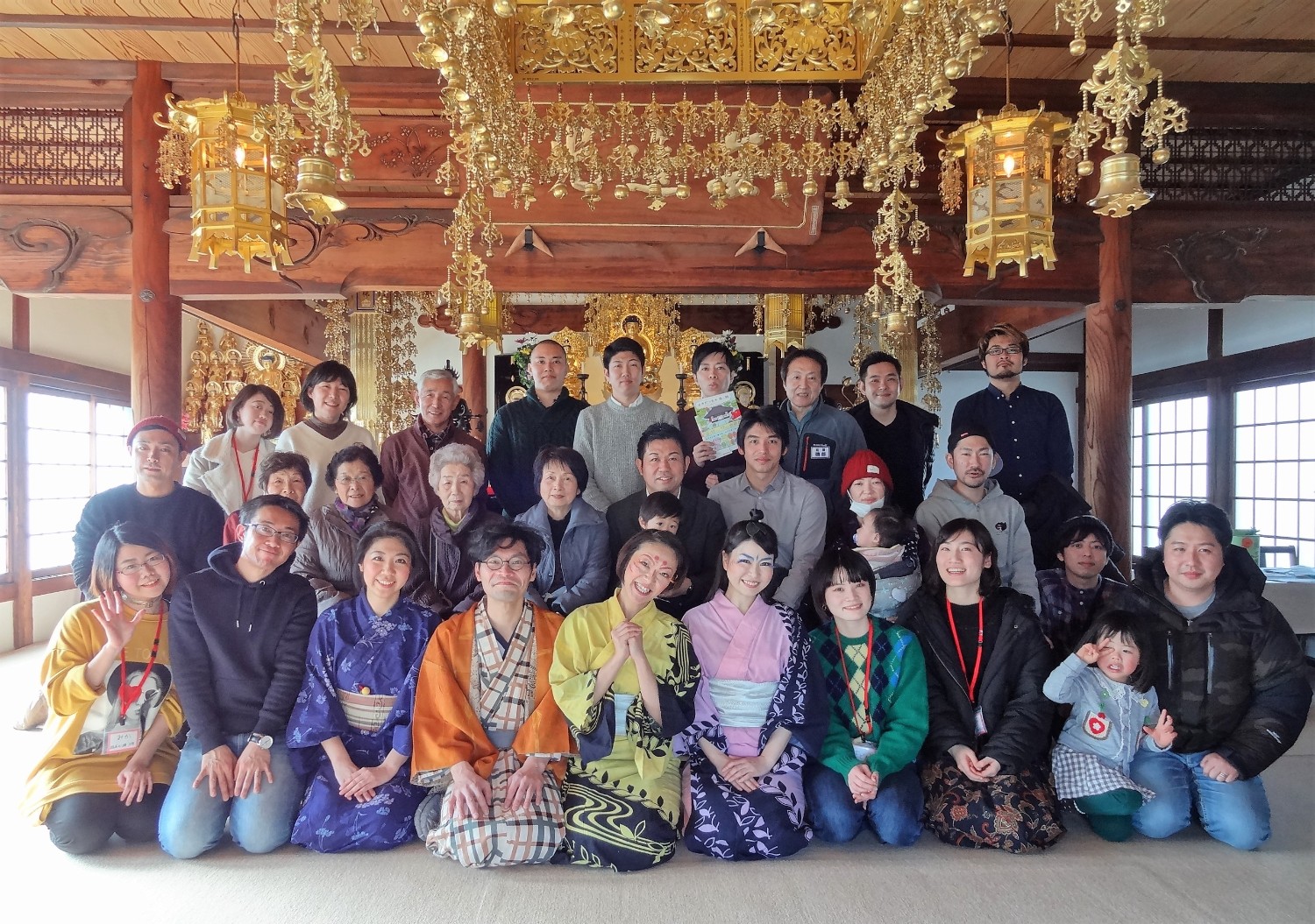 和室の中に金色の装飾が施された豪華な仏壇が置かれた本堂で、ごきげん一家のメンバーとスタッフ、観客・来場者で記念撮影を行っている写真