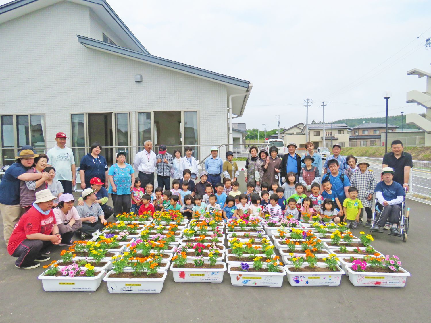 白い建物の前にきれいに並んだ花を植えた沢山の白いプランターの周りに、多くの子どもたちと高齢者を含む大人たちが集まっている集合写真