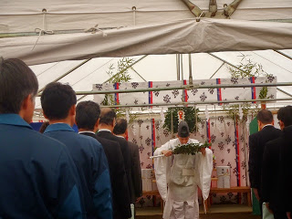 出席者たちが席から立ち上がり起立し、前方では神職の男性が榊を手に礼をしている様子の写真