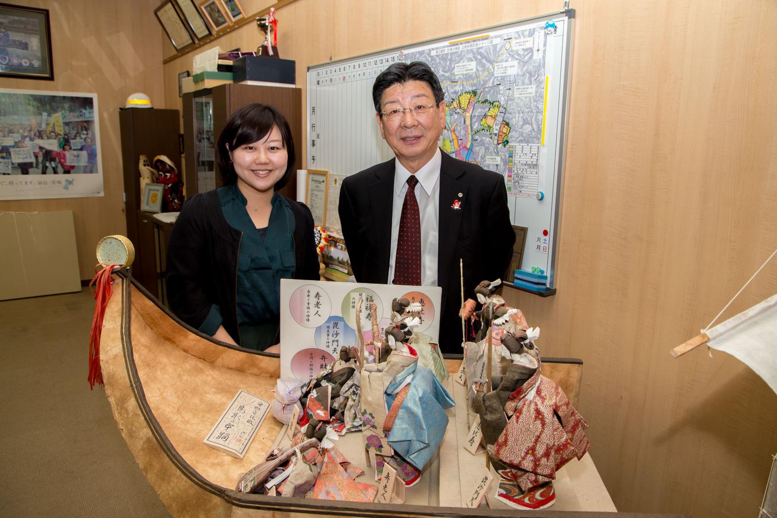 船のような台座に着物を着た日本人形が数体並んでいる飾りの後ろに笑顔で立っている女性リポーターと佐藤町長の写真