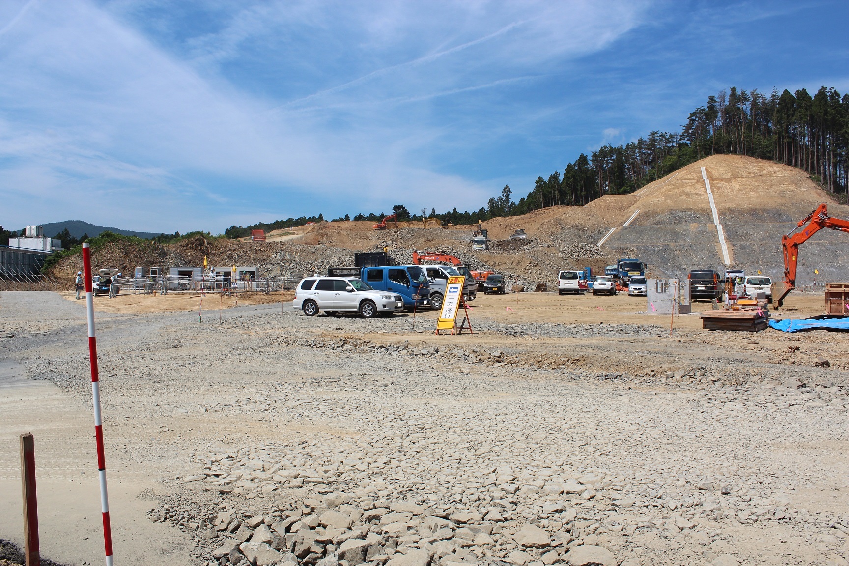 青空の下、まだ整地の途中段階にある広大な造成地に重機や工事車両が多数並び、背後の丘陵地には白いラインで区画された法面が見える造成工事中の様子を捉えた写真