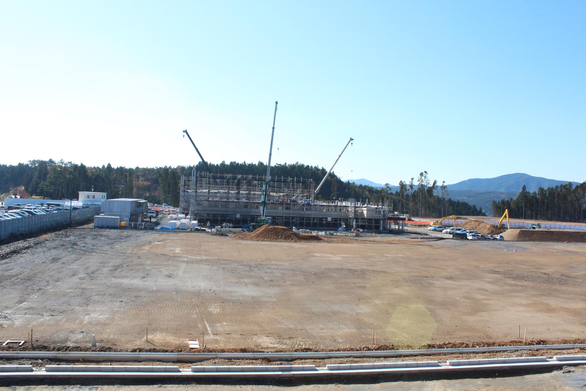 造成された広大な敷地の奥に足場に囲まれた鉄骨建築の建物が立ち上がり、3台のクレーンが建設作業を行っている様子の写真