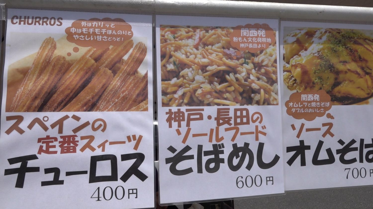 白い屋台テントの下に並べられた、左からチュロス、そばめし、ソースオムそばの料理写真と価格が大きく印刷されたメニューの貼り紙の写真