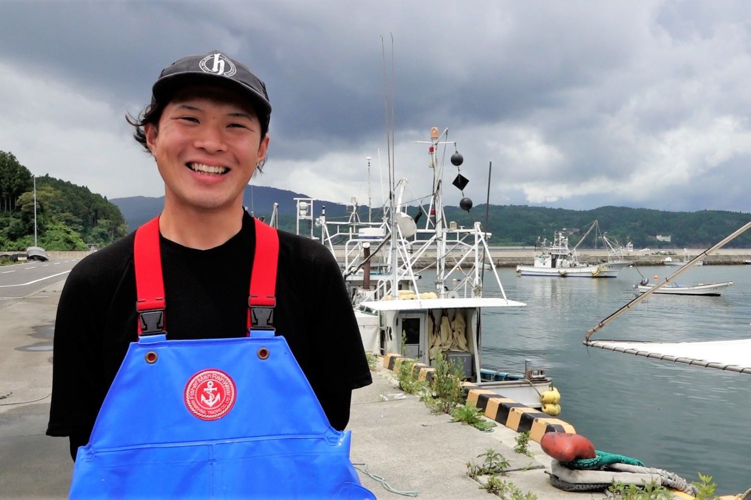 港に停泊した漁船を背景に、青い前掛けと黒いシャツ姿の若い男性が防波堤の上で満面の笑顔を見せて立っており、曇り空の下で活気ある港町の雰囲気が伝わる写真